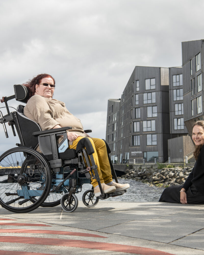 Lady with red hair sitting in a netti v side on, with her facelooking at the camera and her friend sat beside her on the concrete floor