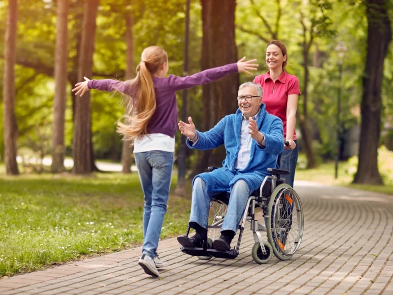 A man being pushed in a wheelchair by a yound lady with his hands our to greeet a young girl running towards him