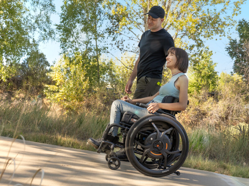 A young lady on a path sittin in a wheelchair with m90 wheels and a man walkng alongside her