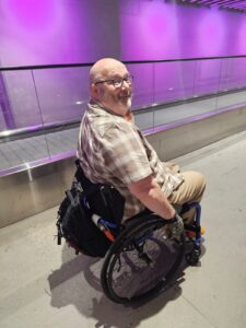 MAN WITH HIS FACE TURNED TOWARDS THE CAMERA SAT IN A REHASENSE BLUE WHEELCHAIR ON AN ACCESS RAMP INDOORS