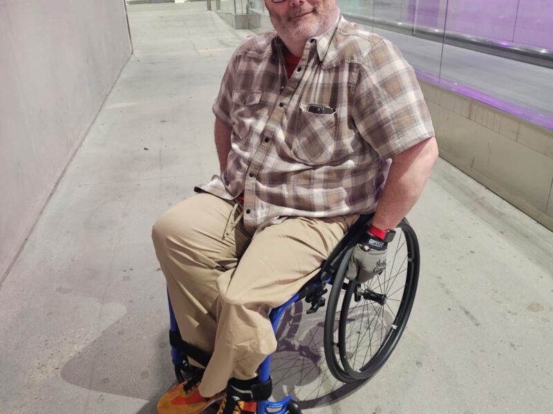 MAN SAT FACING THE CAMERA IN A BLUE ICON WHEELCHAIR ON A RAMP INDOORS