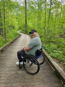 MAN SAT IN AN ICON WHEELCHAIR WITH HIS FACE LENT TOWARDS THE CAMERA ON A BOARDWALK IN A WOODED AREA